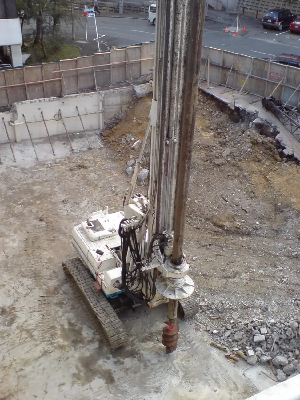 A piling rig drilling with an auger in a city-centre basement dig, behind a concrete retaining wall