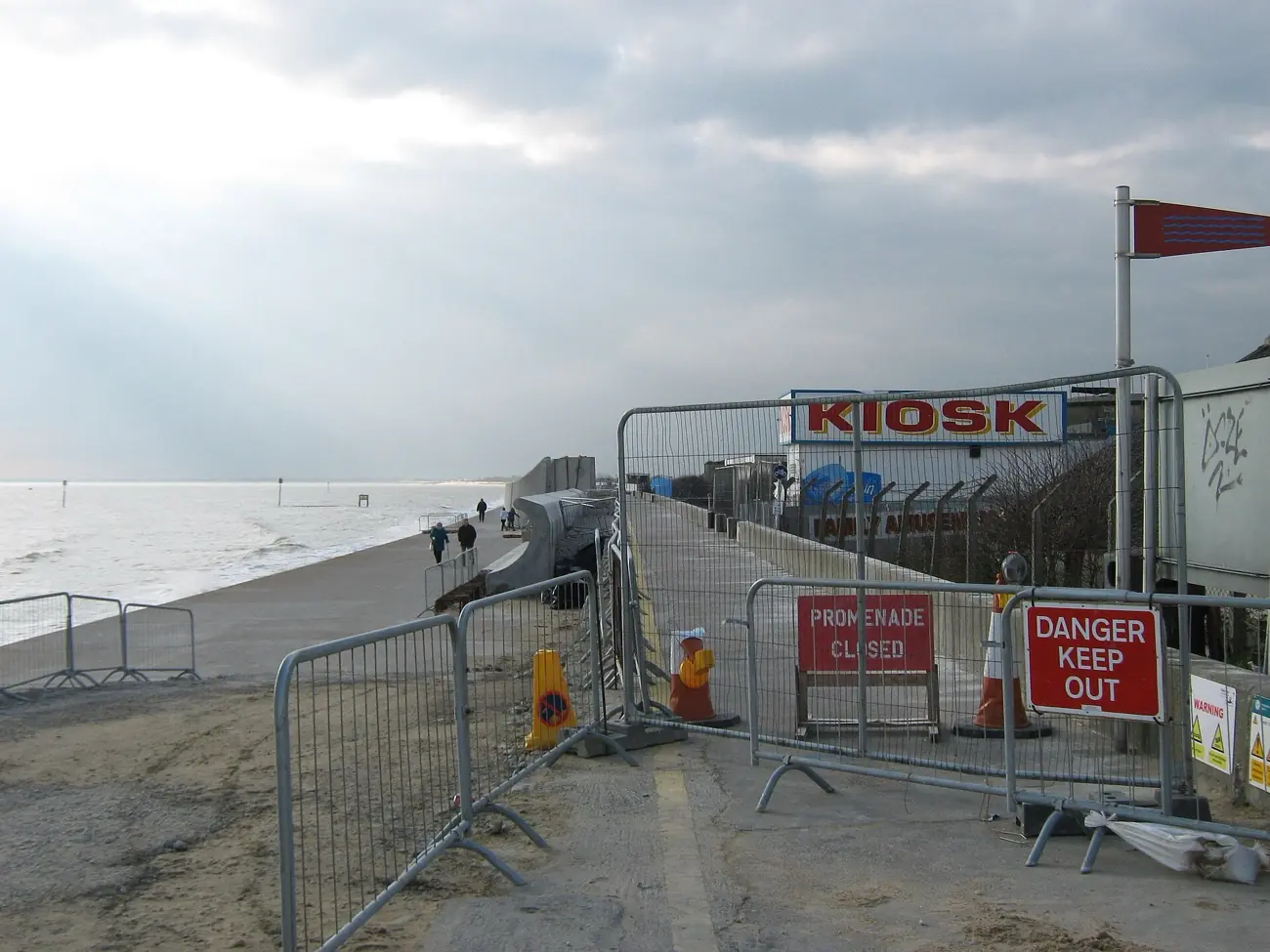 A closed seafront promenade behind site fencing, with the sea wall works beyond